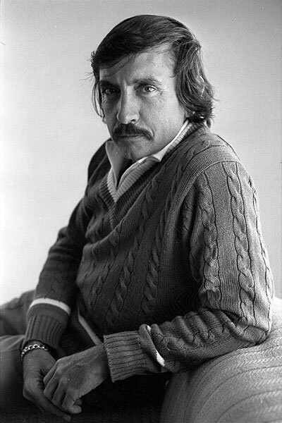 A man with light skin, medium length hair and a moustache, wearing a cable-knit sweater and leaning on a couch.