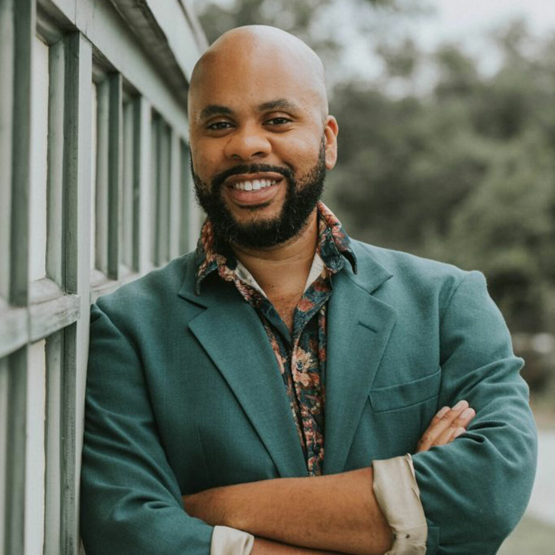 A bald, bearded, smiling man with brown skin wearing a teal blazer and floral-print shirt, leaning against a building with his arms crossed.