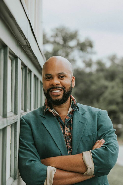 A bald, bearded, smiling man with brown skin wearing a teal blazer and floral-print shirt, leaning against a building with his arms crossed.