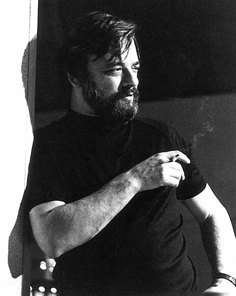 A bearded man in a black t-shirt stands in a dramatic pose holding a cigarette in one hand.