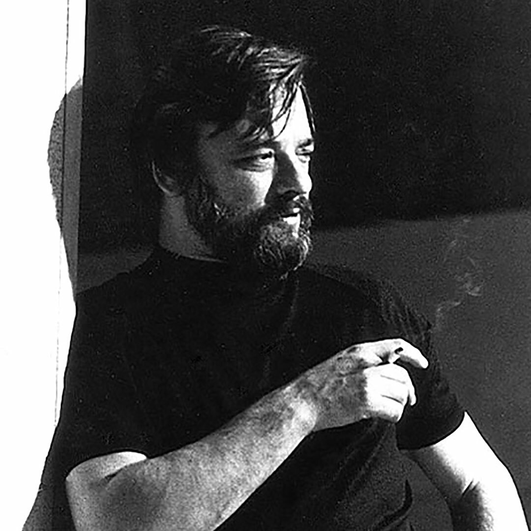 A bearded man in a black t-shirt stands in a dramatic pose holding a cigarette in one hand.