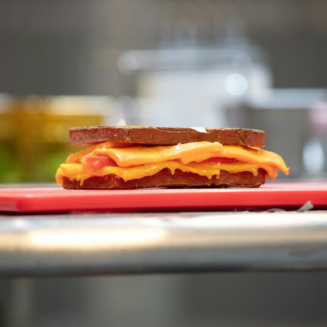 A prop sandwich with realistic bread and melted cheese slices resting atop a red cutting board on a metal table.