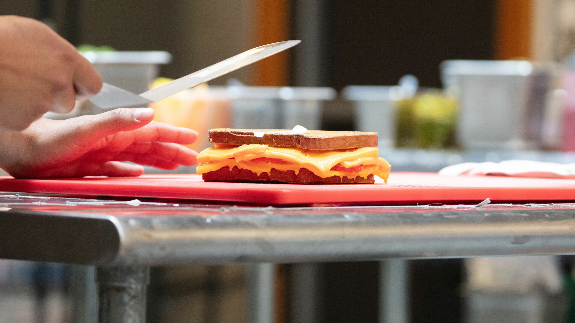 Hands hold a spatula over a prop sandwich with realistic bread and melted cheese slices resting atop a red cutting board on a metal table.