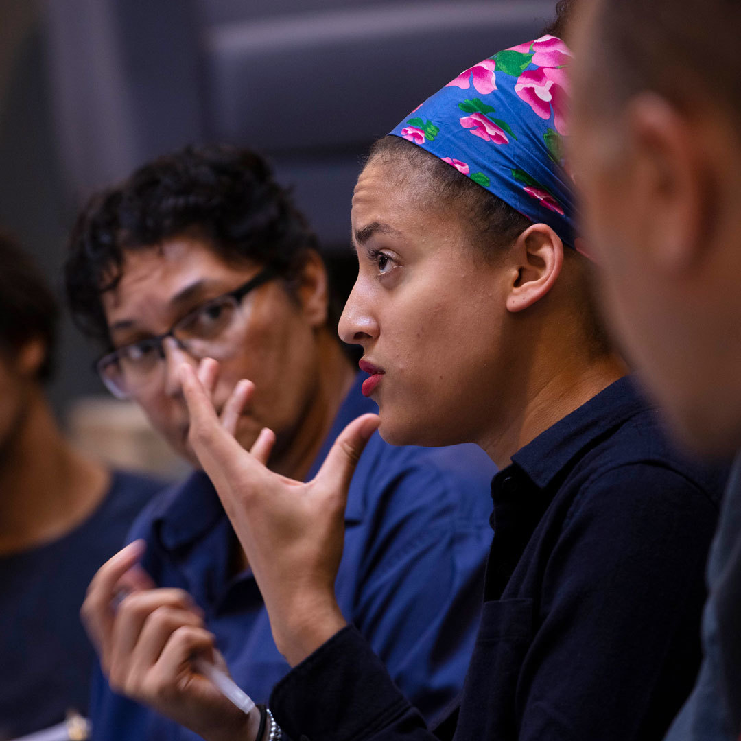 A person of color is speaking and gesturing with their hands as three other people look on.