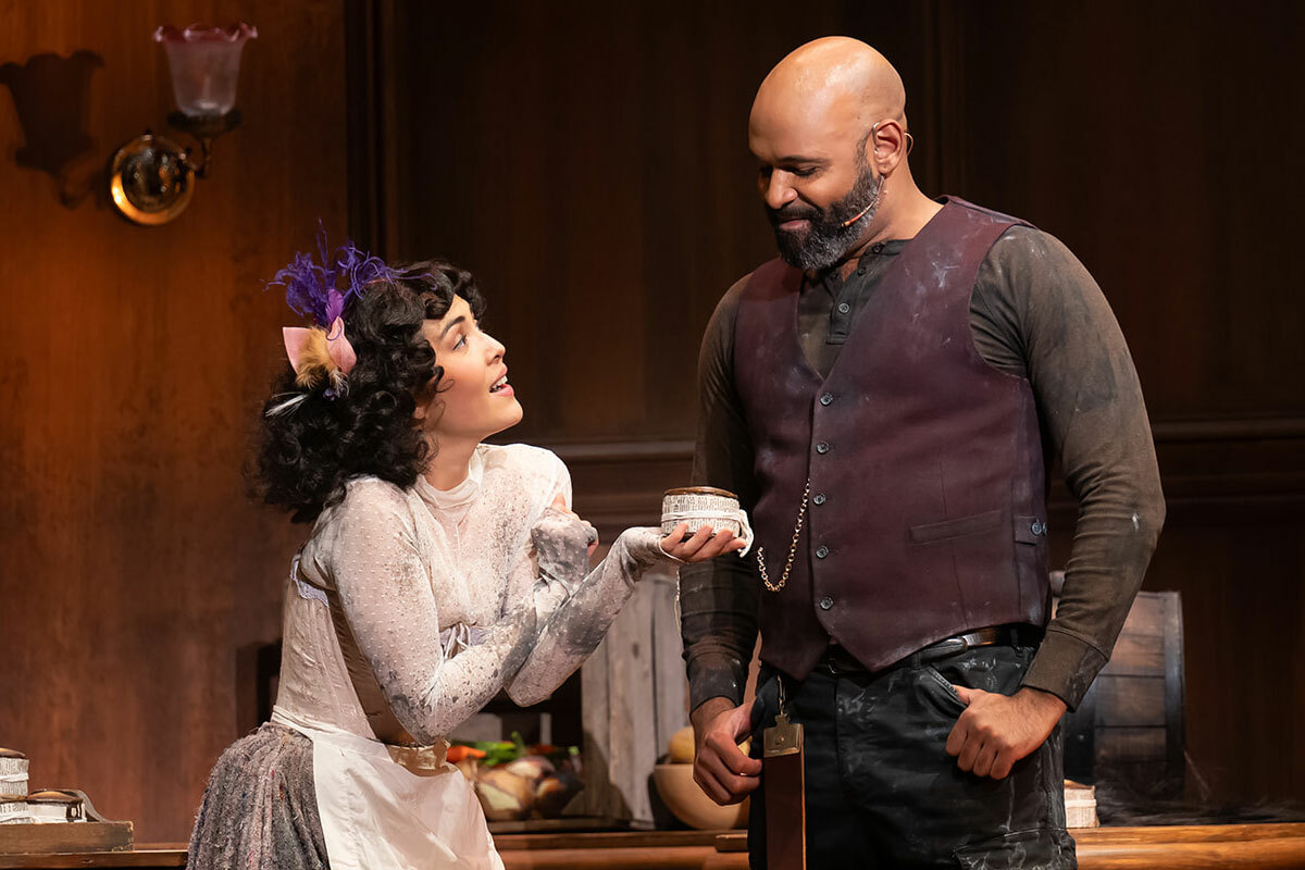 Two characters on stage: a dark-haired woman in a white dress and apron offers a meat pie to a bald, bearded man wearing a vest.