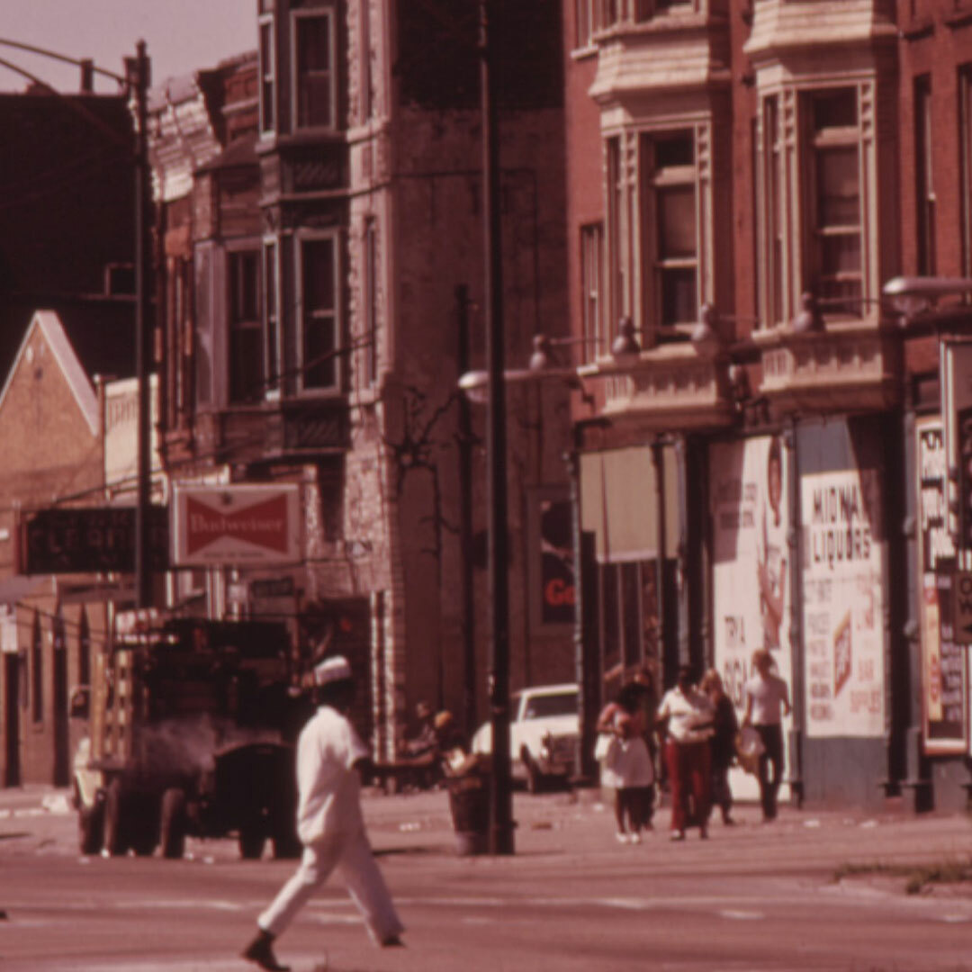 An urban street lined with shops, apartment buildings, cars, people on the sidewalk, and a person crossing in the foreground.