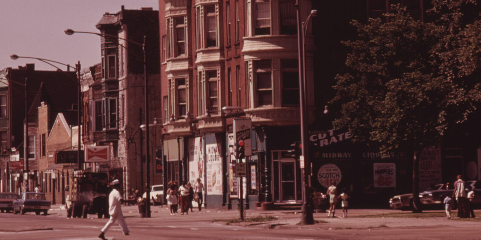 An urban street lined with shops, apartment buildings, cars, people on the sidewalk, and a person crossing in the foreground.