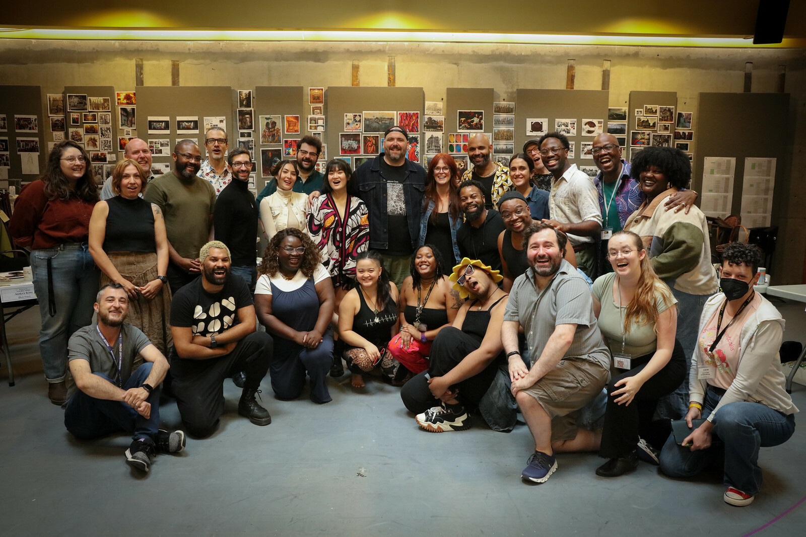 A group of 28 people posed in standing and seated positions in a theater rehearsal room.