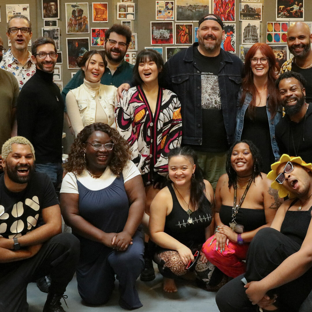 A group of 28 people posed in standing and seated positions in a theater rehearsal room.